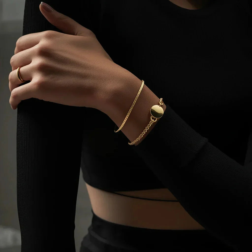 A model wearing a layered gold bracelet set by TM & Co. Jewels, featuring a sleek serpentine chain and a thick curb chain with a circular charm.