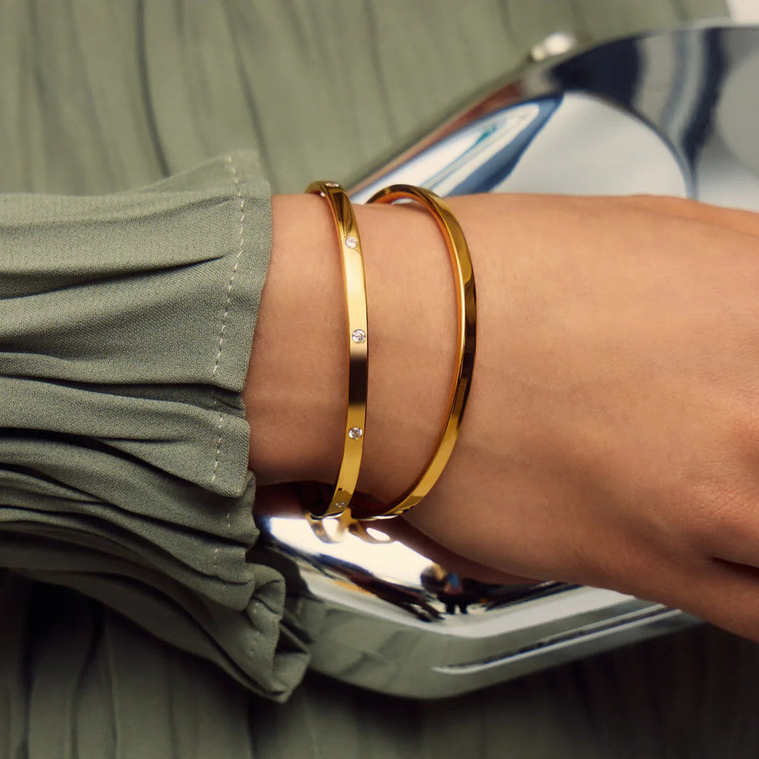Gold bangles on a person's wrist with a green jacket and reflective surface in the background