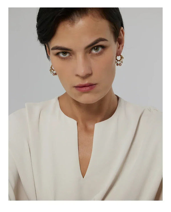 Woman wearing gold-plated dangle earrings with gemstones, cream blouse, neutral background