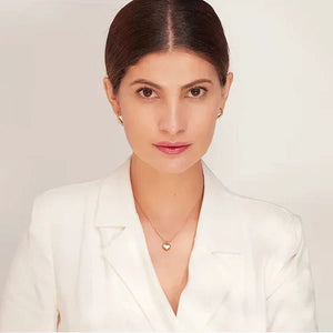 A woman with dark hair pulled back wears a white blazer and a delicate gold heart pendant necklace against a clean off-white background.