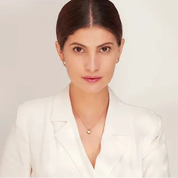 A woman with dark hair pulled back wears a white blazer and a delicate gold heart pendant necklace against a clean off-white background.