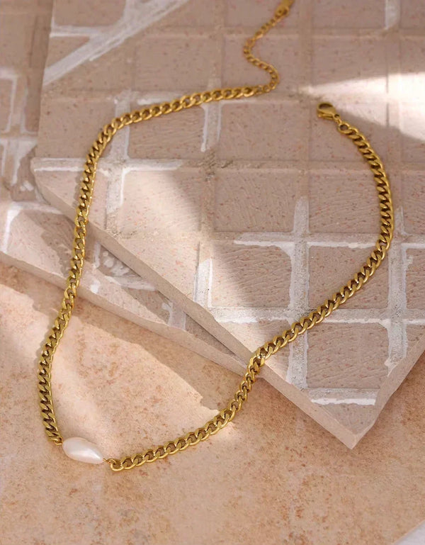 A gold chain necklace with a small white pearl pendant, styled on a beige stone tile background.