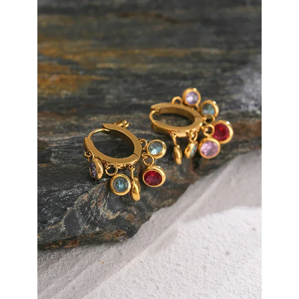Gold-plated hoop earrings with colorful gemstone charms on a stone background