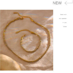 Gold necklace and bracelet set on a textured beige surface with 'NEW' label.