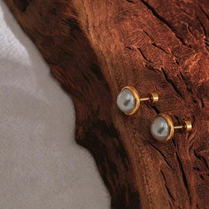 Gold-plated pearl stud earrings on textured wood and fabric background