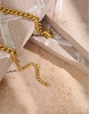 A close-up of a gold chain necklace with a curb link style and lobster clasp, draped over layered beige stone blocks.