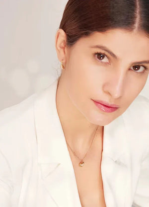 A close-up portrait of a woman wearing a delicate gold necklace with a small gold pendant and a matching small gold hoop earring, paired with a white blazer against a soft light gray background.