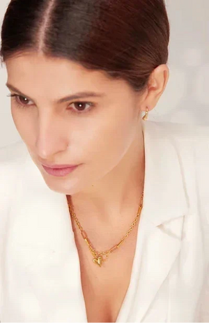 A close-up of a woman wearing a delicate gold chain necklace with a small heart-shaped pendant, styled with a matching small gold hoop earring, against a clean white blazer and light background.