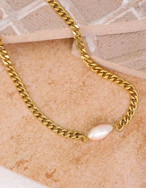 A gold curb chain necklace featuring a single baroque pearl pendant, displayed on a textured beige surface.
