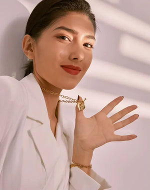 A woman wearing a white blazer and gold jewelry, including a gold chain necklace with a heart-shaped pendant and a gold bracelet, posing with her hand raised against a soft, light-colored background with shadow patterns.