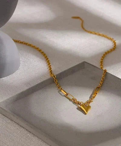 A gold chain necklace with a small heart-shaped pendant, displayed on a light gray textured tray against a white background with soft shadows.