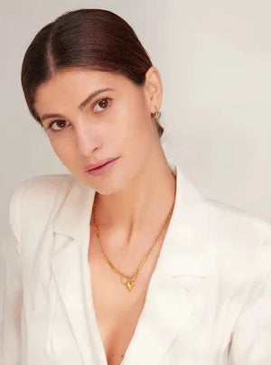 A woman with dark hair pulled back wears a white blazer and a delicate gold necklace with a small heart-shaped pendant, photographed against a light neutral background.