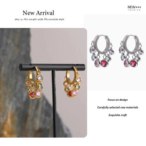 Pair of hoop earrings with gemstones on a stand against a neutral background, with text indicating 'New Arrival' and brand name.