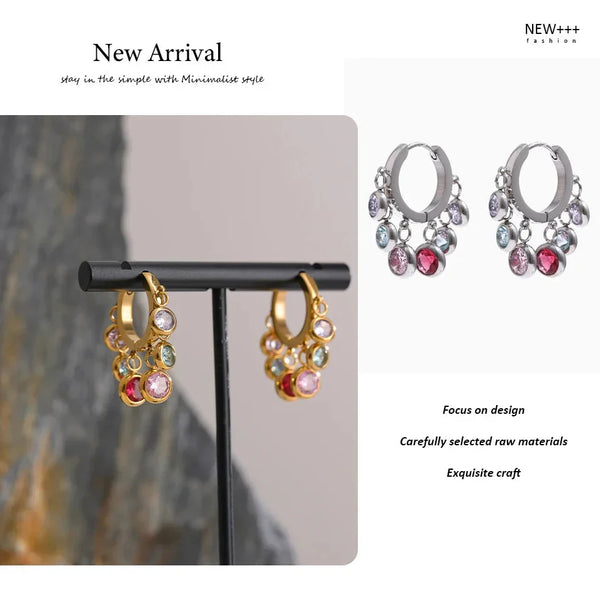 Pair of hoop earrings with gemstones on a stand against a neutral background, with text indicating 'New Arrival' and brand name.