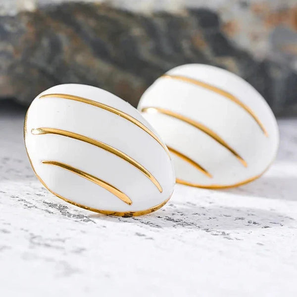 A pair of white oval stud earrings with gold curved stripe detailing, resting on a light textured surface with a blurred gray rock in the background.