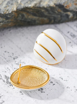 A pair of gold and white enamel egg-shaped earrings with a hammered gold texture and gold stripes, displayed on a light stone surface with a dark rock in the background.