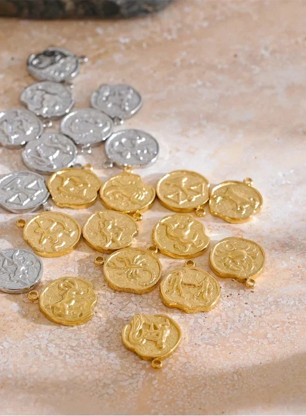 A group of small round coin-shaped charms or pendants, including gold and silver pieces, arranged on a textured beige surface.