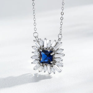 A silver-tone necklace featuring a central square-cut deep blue gemstone surrounded by a halo of clear baguette and round-cut stones, suspended from a delicate silver chain against a light gray background.
