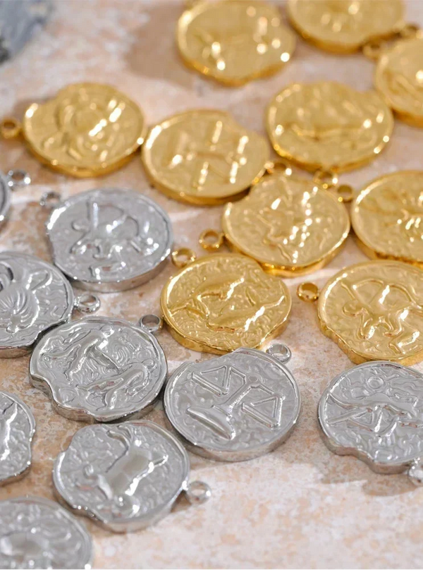 A close-up of small round pendant charms or coins with embossed symbols, shown in both gold and silver finishes scattered on a light stone-like surface.