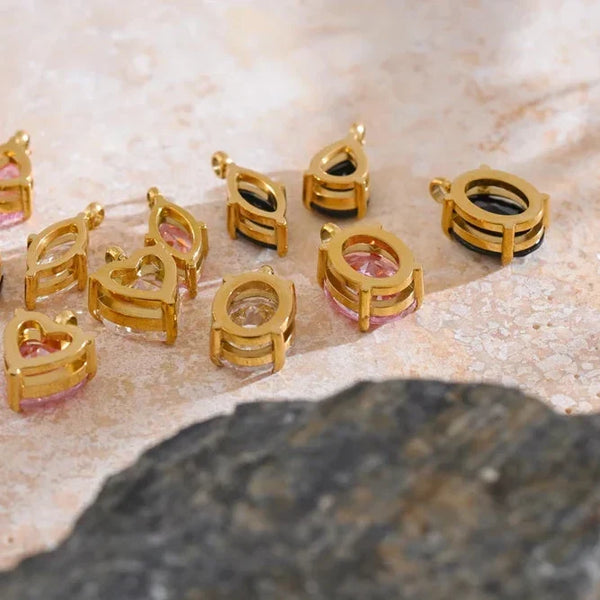 A group of gold-toned heart-shaped jewelry charms or pendants with loop tops, arranged on a light beige stone surface with a dark rock in the foreground.