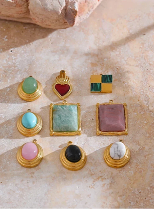 Nine gold jewelry pieces with various colored stones are arranged on a textured beige stone surface. The collection includes oval and square bezel settings in green, turquoise, pink, black, white, and red, alongside a heart-shaped pendant and a small stacked green-and-gold square charm.