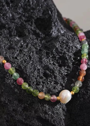 A beaded necklace featuring a mix of colorful round gemstone beads in pink, green, orange, and yellow, accented with small gold spacers and a large white pearl bead, displayed on a textured black rock.
