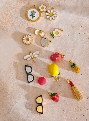 A collection of colorful enamel jewelry charms and earrings with gold-tone borders, arranged on a textured beige stone surface. The charms include daisies, a flower with a purple flower, a duck, a dragonfly, strawberries, a pineapple, a lemon, a pink fruit, a lipstick tube, and a pair of black-rimmed glasses.