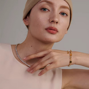 A close-up lifestyle shot of a model wearing a delicate silver tennis necklace, a gold beaded bracelet, and a gold ring, paired with a beige beanie and a pale pink top against a neutral gray background.