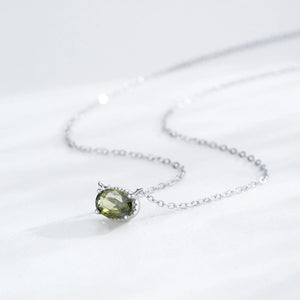 A delicate silver chain necklace featuring an oval-cut olive green gemstone pendant in a silver bezel setting, displayed on a soft white fabric background.