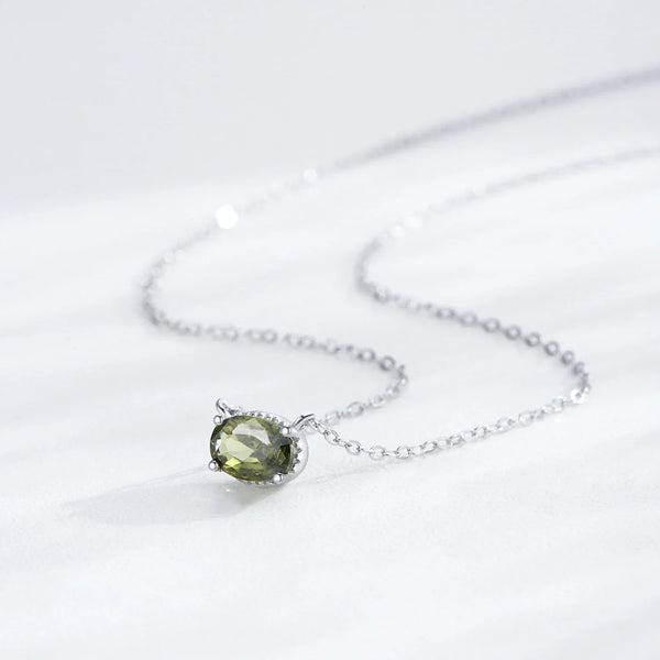 A delicate silver chain necklace featuring an oval-cut olive green gemstone pendant in a silver bezel setting, displayed on a soft white fabric background.