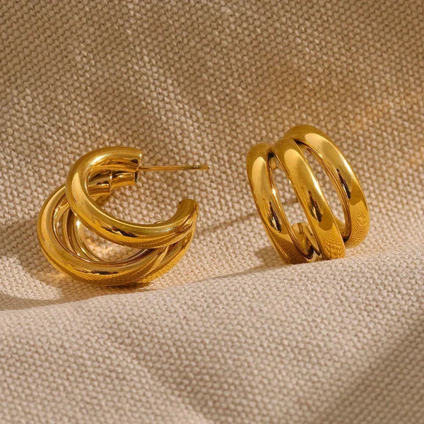Chunky gold-plated hoop earrings with a triple band design on beige textured fabric.