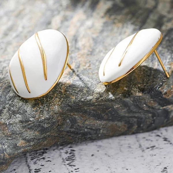 A pair of white oval stud earrings with gold-toned metal edges and three vertical gold lines, displayed on a textured gray stone surface.