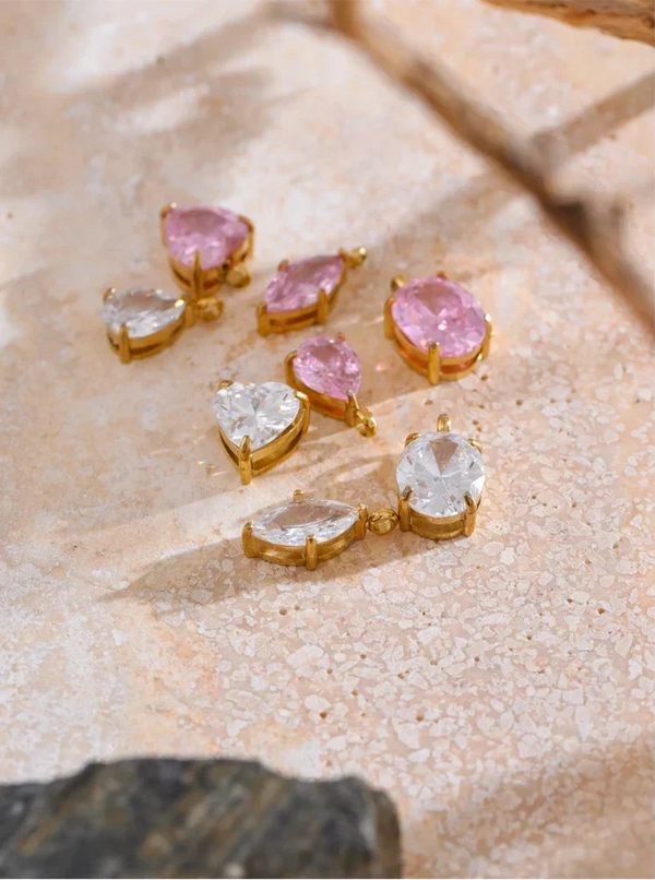 Six gold-toned jewelry findings or earrings with pink and clear stones, arranged on a textured beige stone surface.