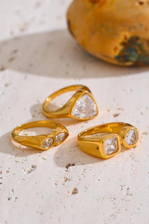 Three gold rings with clear gemstones on a textured beige surface.