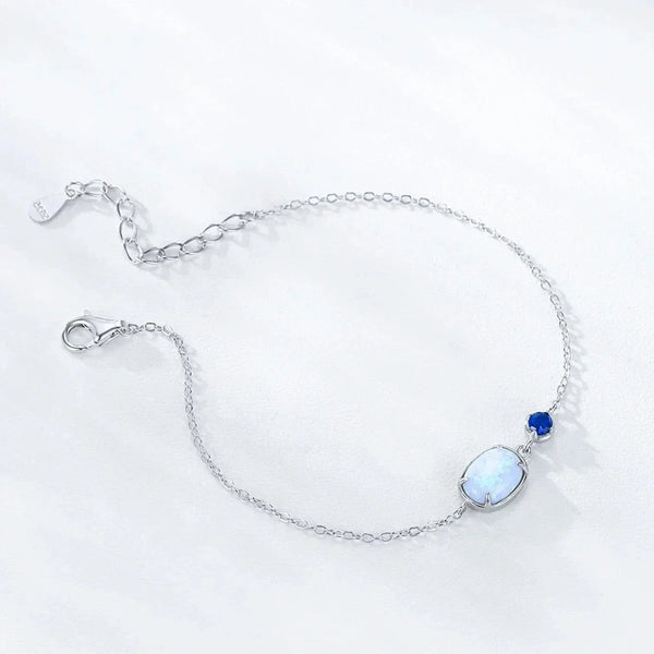 A delicate silver chain bracelet featuring a large oval white opal centerpiece and a small round blue gemstone accent, displayed on a clean white background.