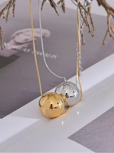 Two spherical pendant necklaces, one gold and one silver, hanging from delicate chains against a styled background with dried branches and a gray-toned print.