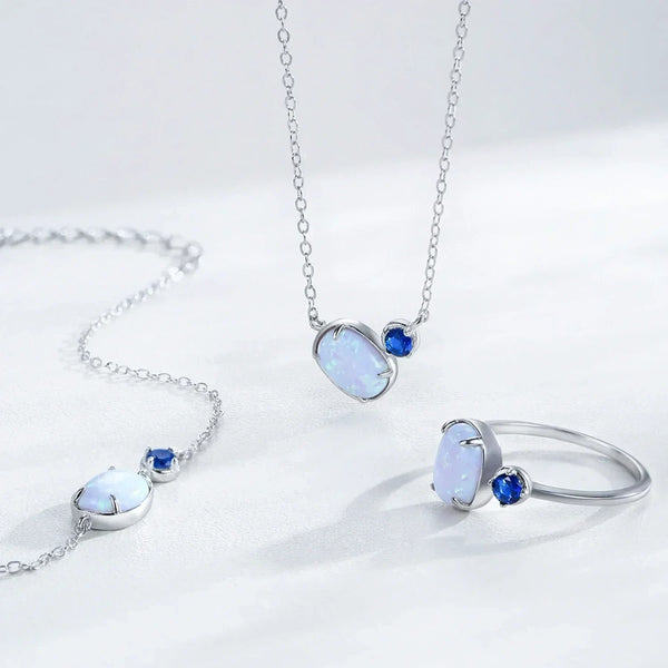 Silver jewelry set featuring a pendant necklace and matching ring, each with an oval white opal-like stone and a small round blue accent stone, displayed on a clean white background.