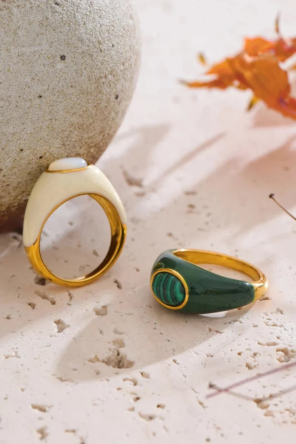 Two rings, one beige and gold, one green and gold, on a textured surface with a blurred natural background.