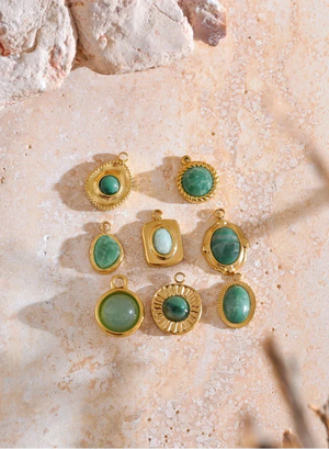 Nine gold jewelry pendants with green stone cabochons in various shapes, arranged on a textured beige stone surface.