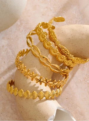 Four gold-toned bangles with intricate floral and shell-like patterns, displayed on a beige stone stand against a soft beige background.
