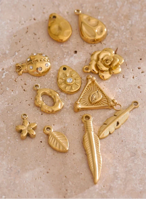 A set of gold-colored metal jewelry charms and pendants arranged on a textured beige stone surface. The charms include a rose, leaves, feathers, a sun face, a flower, a fish, and a crescent moon with small clear rhinestone accents.