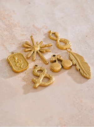 Six gold-toned jewelry charms with bohemian and abstract designs, including a feather, sunburst, infinity, cherries, and abstract symbols, arranged on a textured beige stone surface.