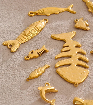 A set of gold-colored fishing lures shaped like fish, fish skeletons, and dolphins, arranged on a textured beige surface.
