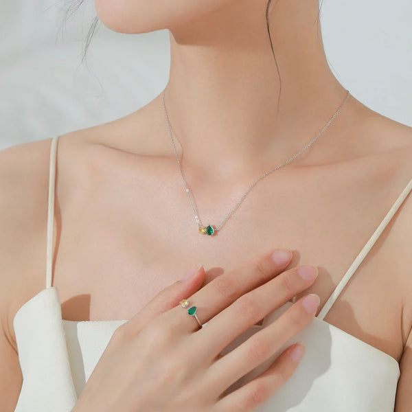 A close-up of a model wearing a delicate silver necklace with a small green and gold pendant, and a matching green ring on her finger, styled with a cream-colored spaghetti strap top.