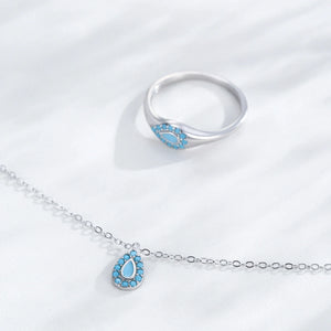 A silver-tone ring and matching chain necklace with a teardrop-shaped pendant, both featuring a light blue inlay surrounded by small clear stones, displayed on a white textured fabric background.