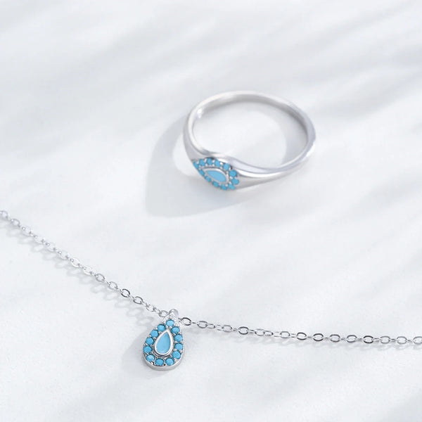 A silver-tone ring and matching chain necklace with a teardrop-shaped pendant, both featuring a light blue inlay surrounded by small clear stones, displayed on a white textured fabric background.