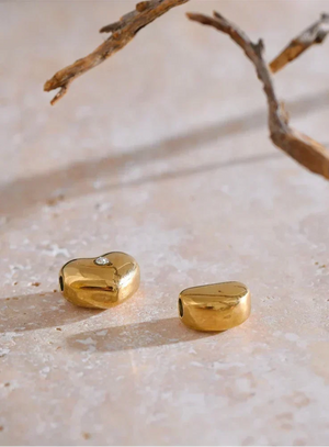 Two small gold-toned jewelry pieces, one heart-shaped and one rounded rectangular, displayed on a light stone surface with a decorative branch overhead.