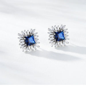 A pair of silver-toned stud earrings with square blue gemstones surrounded by radiating clear crystal-like accents, displayed on a clean white surface with soft shadows. Text at the top reads 'Product Display'.