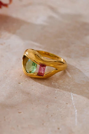 Gold ring with red and green gemstones on a beige surface