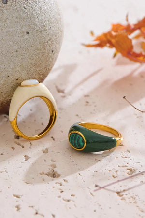 Two rings, one gold and one green, on a textured surface with a stone and leaves in the background.